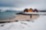 Person walks on a snowy beach near colorful red and yellow cabins. Overcast sky, mountains, and wind turbines in the background.