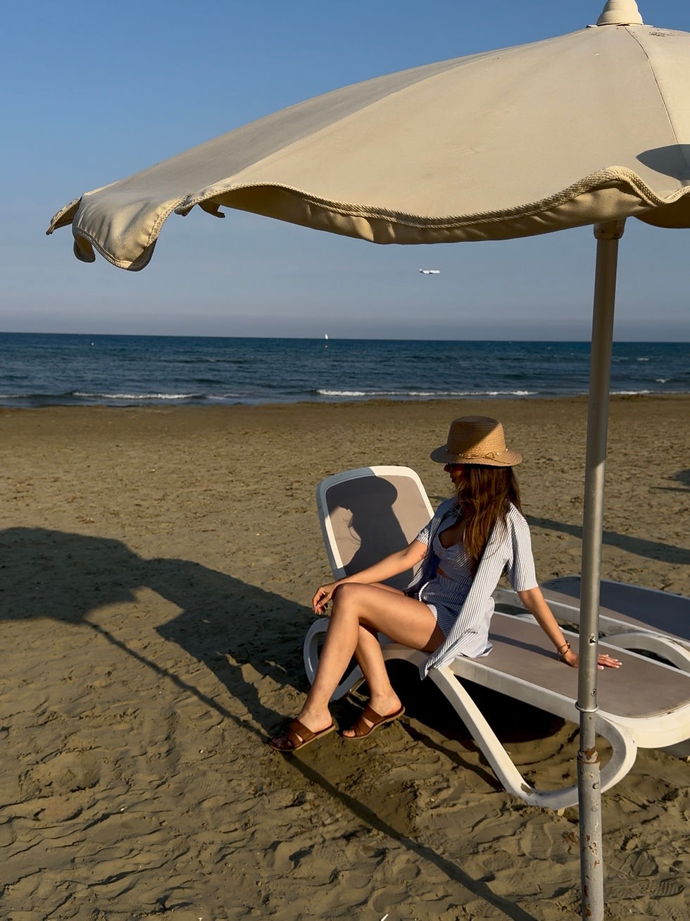 Woman in a straw hat relaxes on a beach chair under a large umbrella, facing the calm sea on a sunny sandy shore.