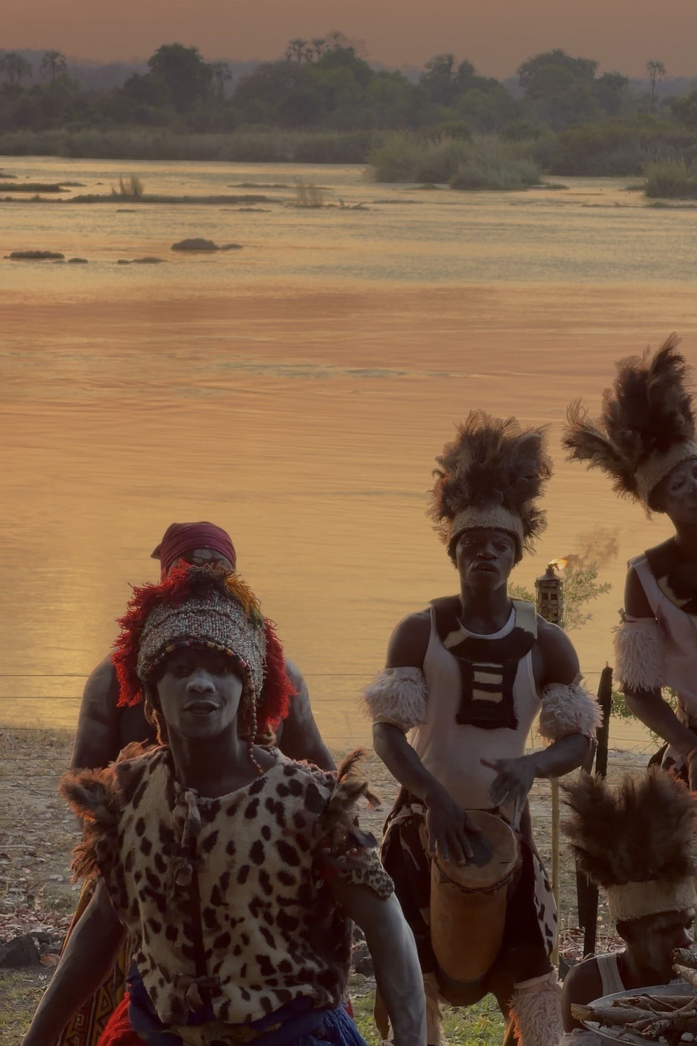 Dancers in animal-print costumes play drums by a serene river at sunset. The scene is vibrant and rhythmic with warm orange tones.