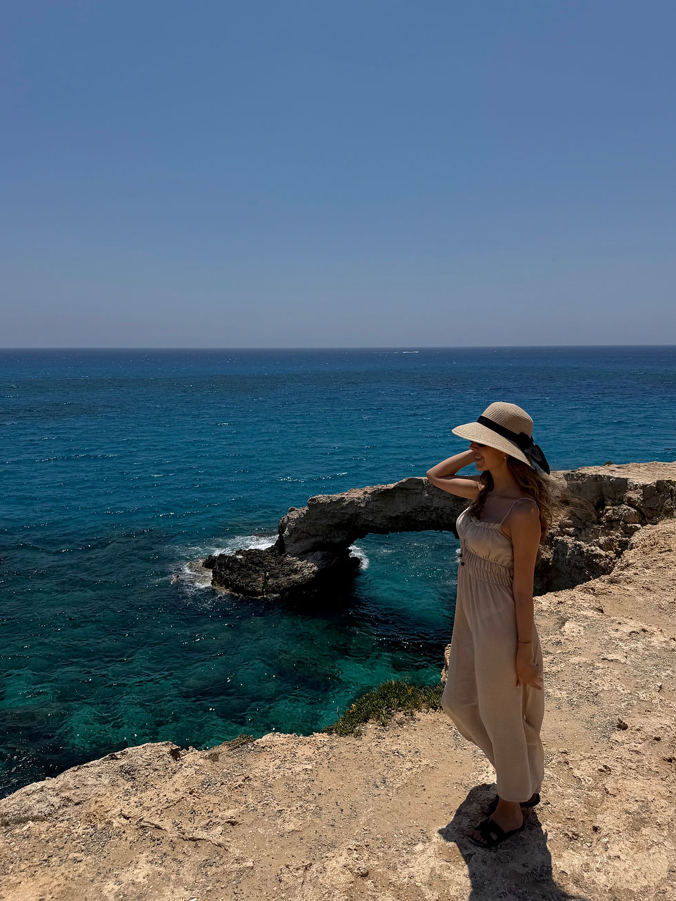 Woman in a straw hat and beige dress stands on a rocky seaside cliff, smiling over the deep blue ocean.
