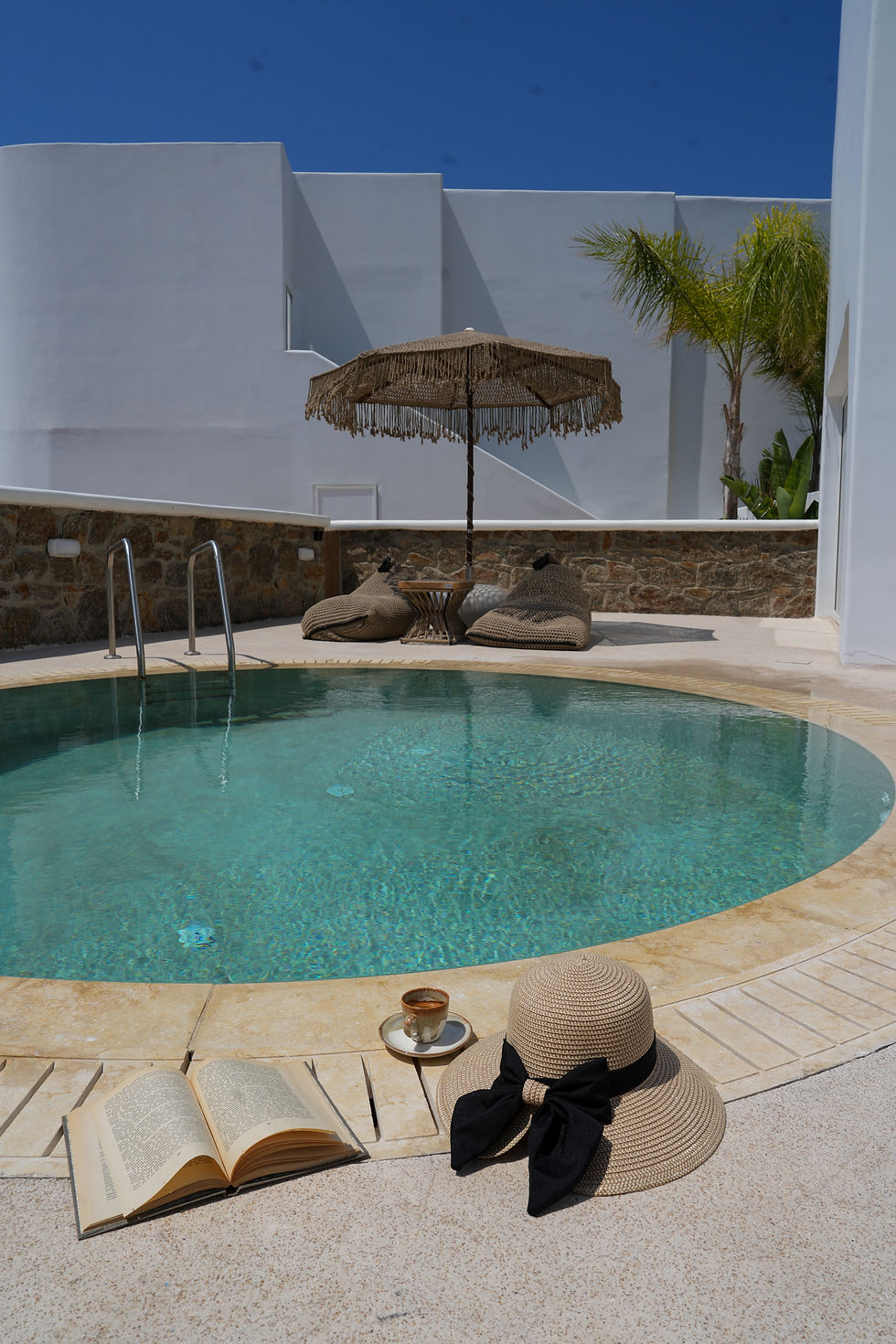 Open book, tea and straw sunhat by a turquoise pool under a fringed umbrella in a quiet sunny courtyard