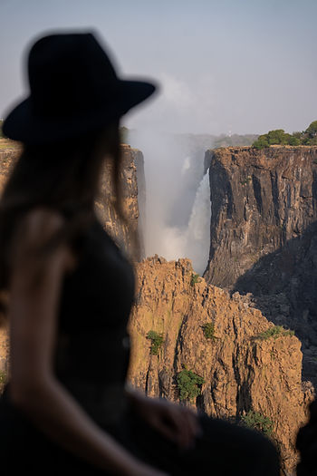 A person in a hat overlooks a waterfall and rocky cliffs under a clear sky. The scene is dramatic and serene.