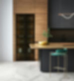 Modern kitchen with wood cabinets, black countertop, and gold bowl. Green bar stool stands on light herringbone floor.