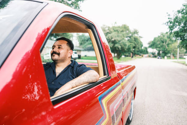 Man smiles while driving a red truck with colorful stripes, parked on a suburban street lined with trees. Bright, relaxed mood.