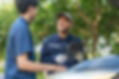 Roadside assistance tech in blue uniform converse outside, one holding a tablet. They're near a car with its hood open, surrounded by green trees.