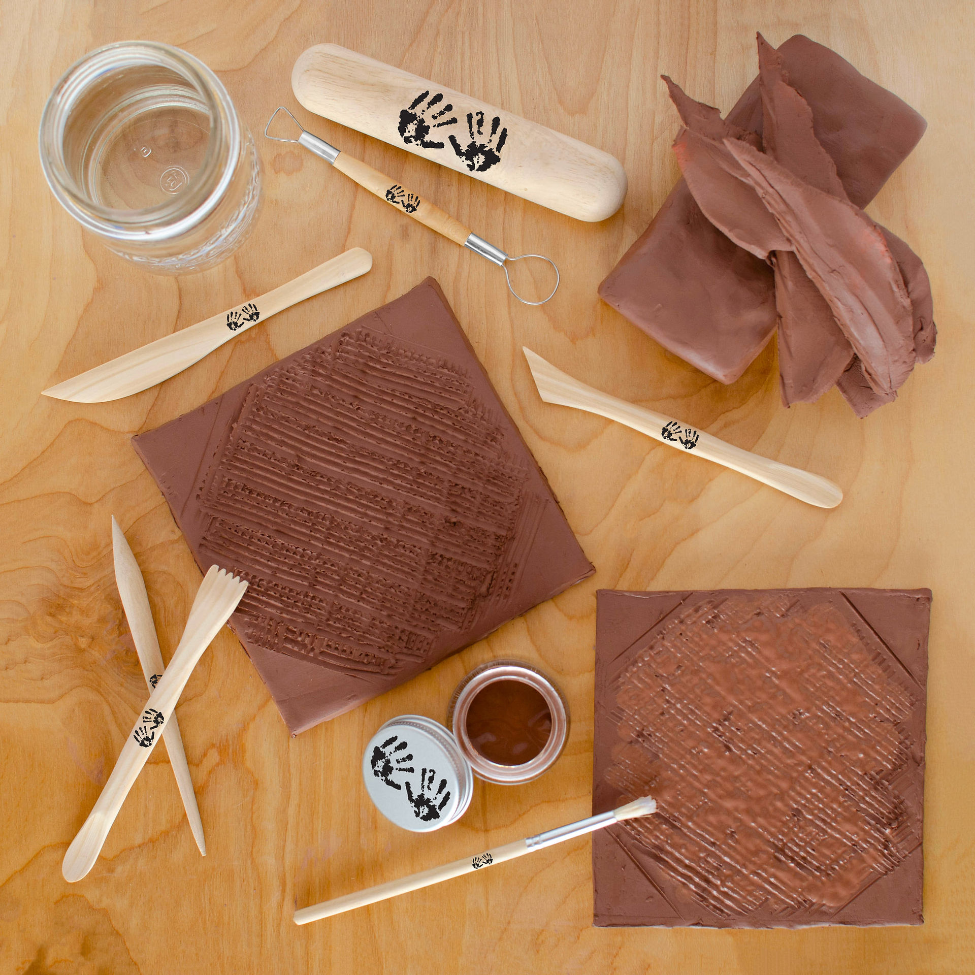 There are clay tools, water, and lumps of terracotta clay on a light wood surface. Two flat squares have been rolled out and trimmed. There are hatch marks on each and a paintbrush is being used to apply slip to each side.