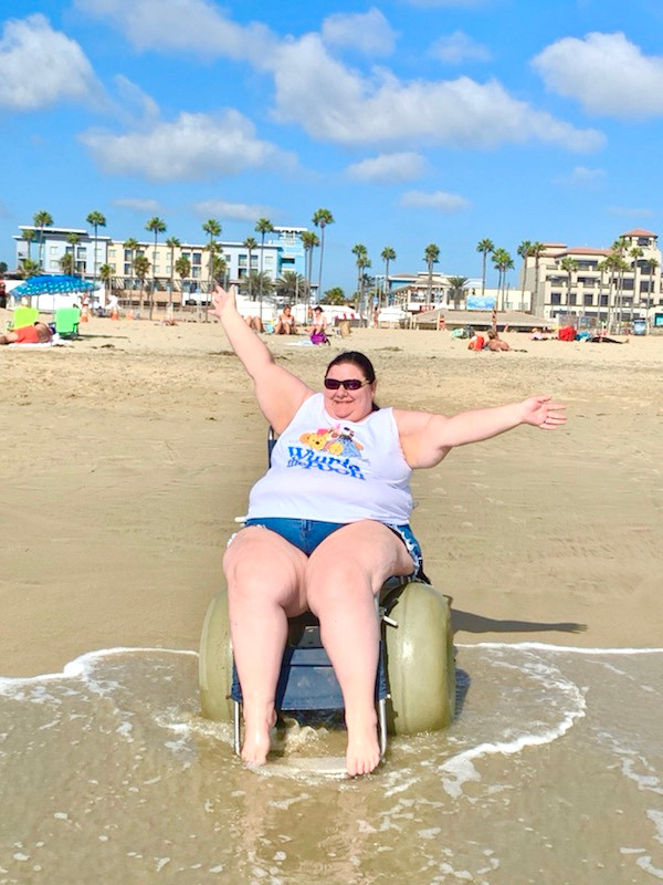 a woman wearing a tank top and jean shorts in a beach wheelchair on the beach