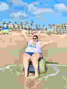 a woman wearing a tank top and jean shorts in a beach wheelchair on the beach