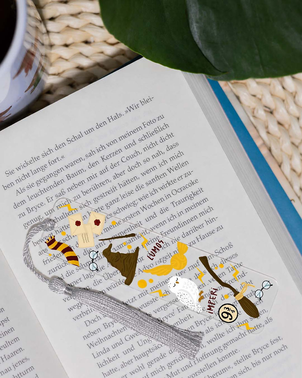 Transparentes Acryl-Lesezeichen mit magischen Zauberwelt-Motiven und silberner Quaste auf einem geöffneten Buch.