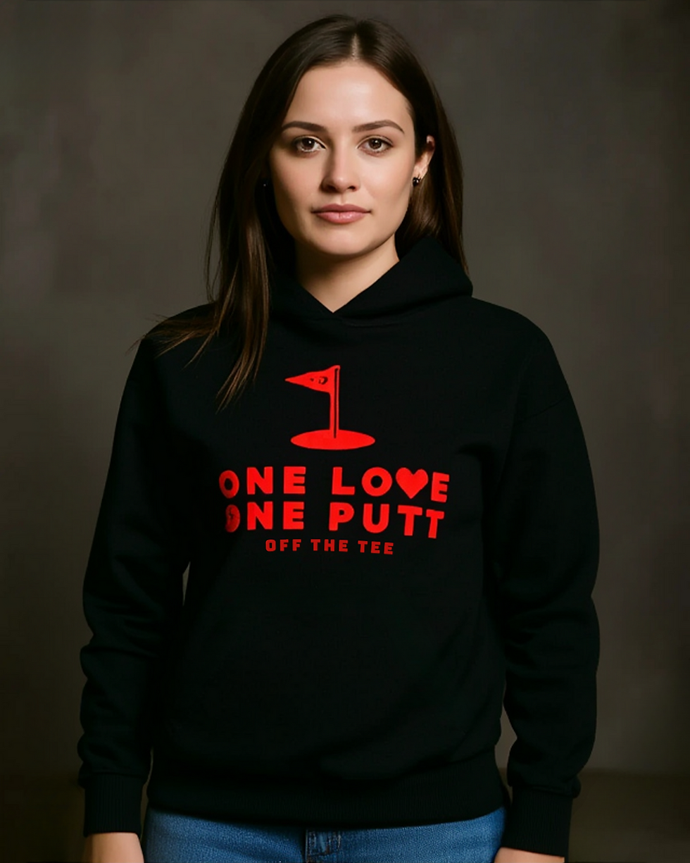 Woman in a black hoodie with red text "ONE LOVE ONE PUTT" and a golf flag design. Neutral expression, plain dark background.
