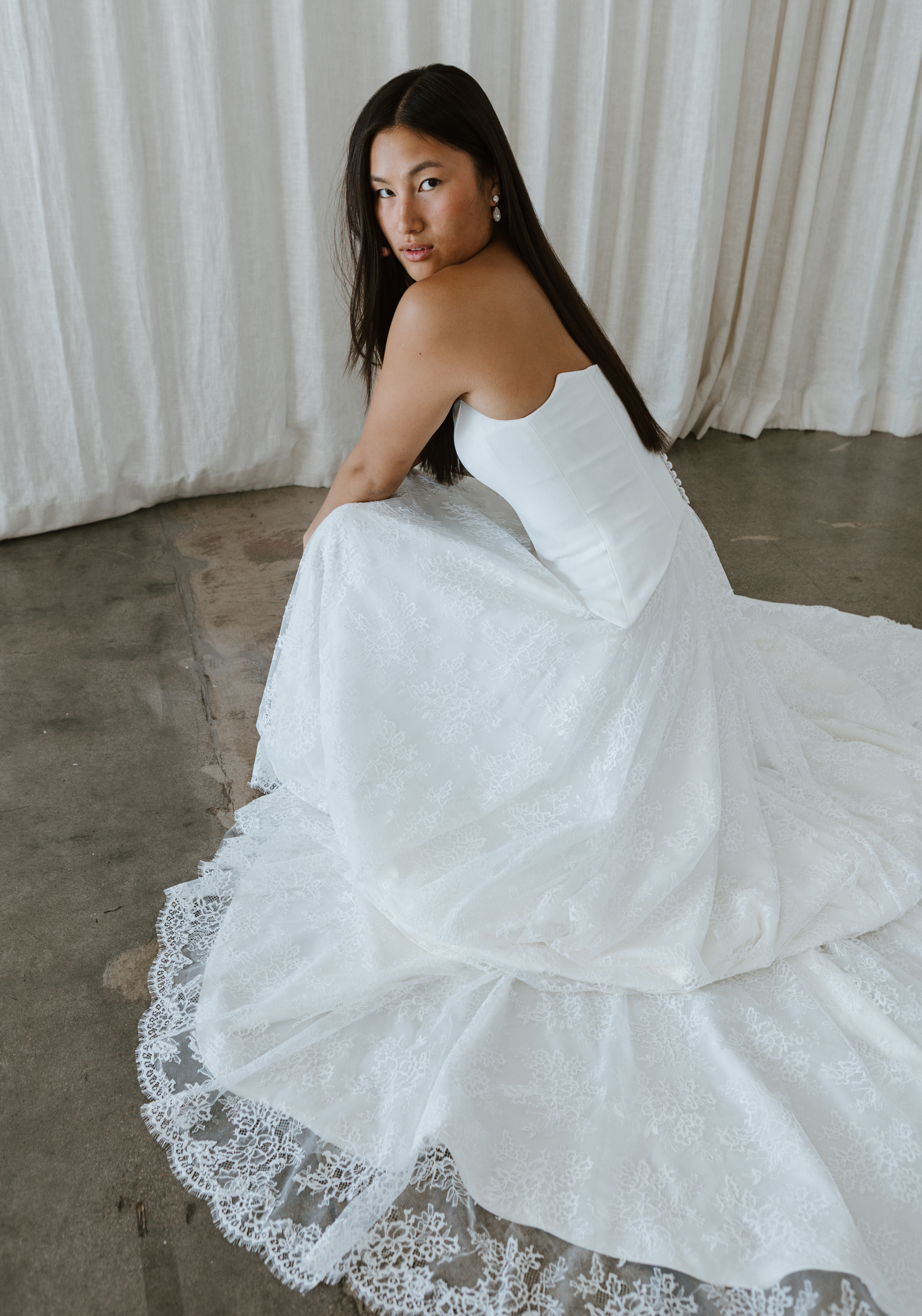 Model is wearing bridal separates in a crouching pose. She's in a strapless, ivory, crepe bodice and her skirt is an a-line cut made in ivory lace.