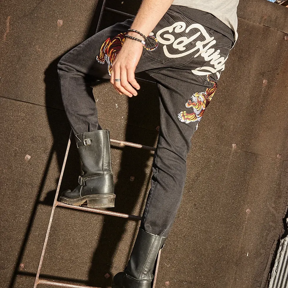 Model wearing black Ed Hardy jeans with bold tiger embroidery and logo script on the back pockets, paired with black leather boots while climbing a metal ladder in an urban setting.