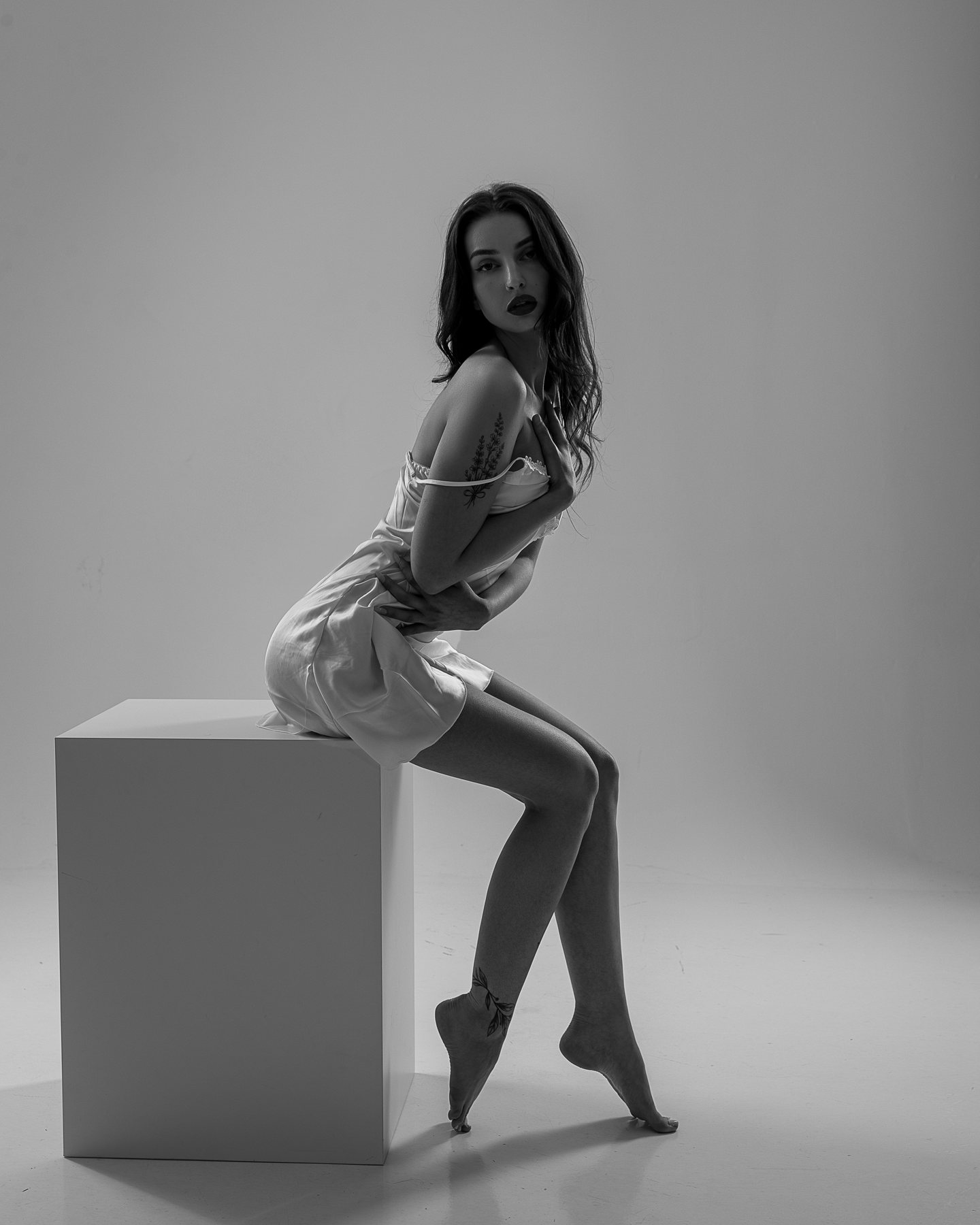 Black and white bridal boudoir portrait of a woman in a white slip seated on a studio block