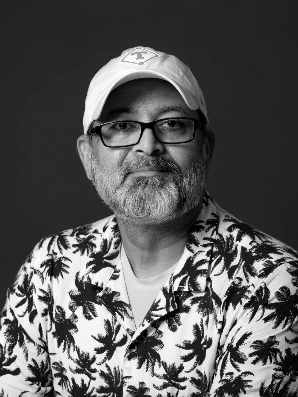Black and white portrait of Mesh Bhakta, Austin boudoir photographer, in a cap and glasses against a dark studio background