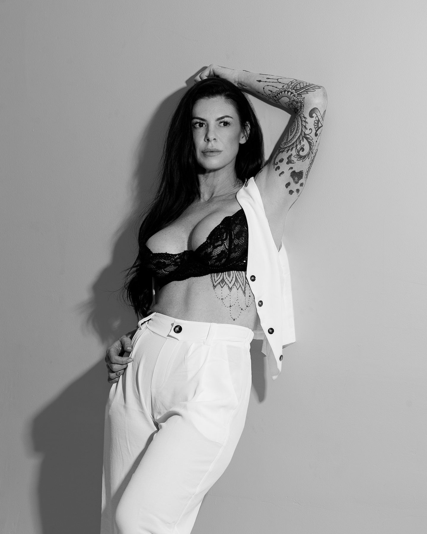 Black and white boudoir portrait of a woman in a black lace bra, white vest and white trousers