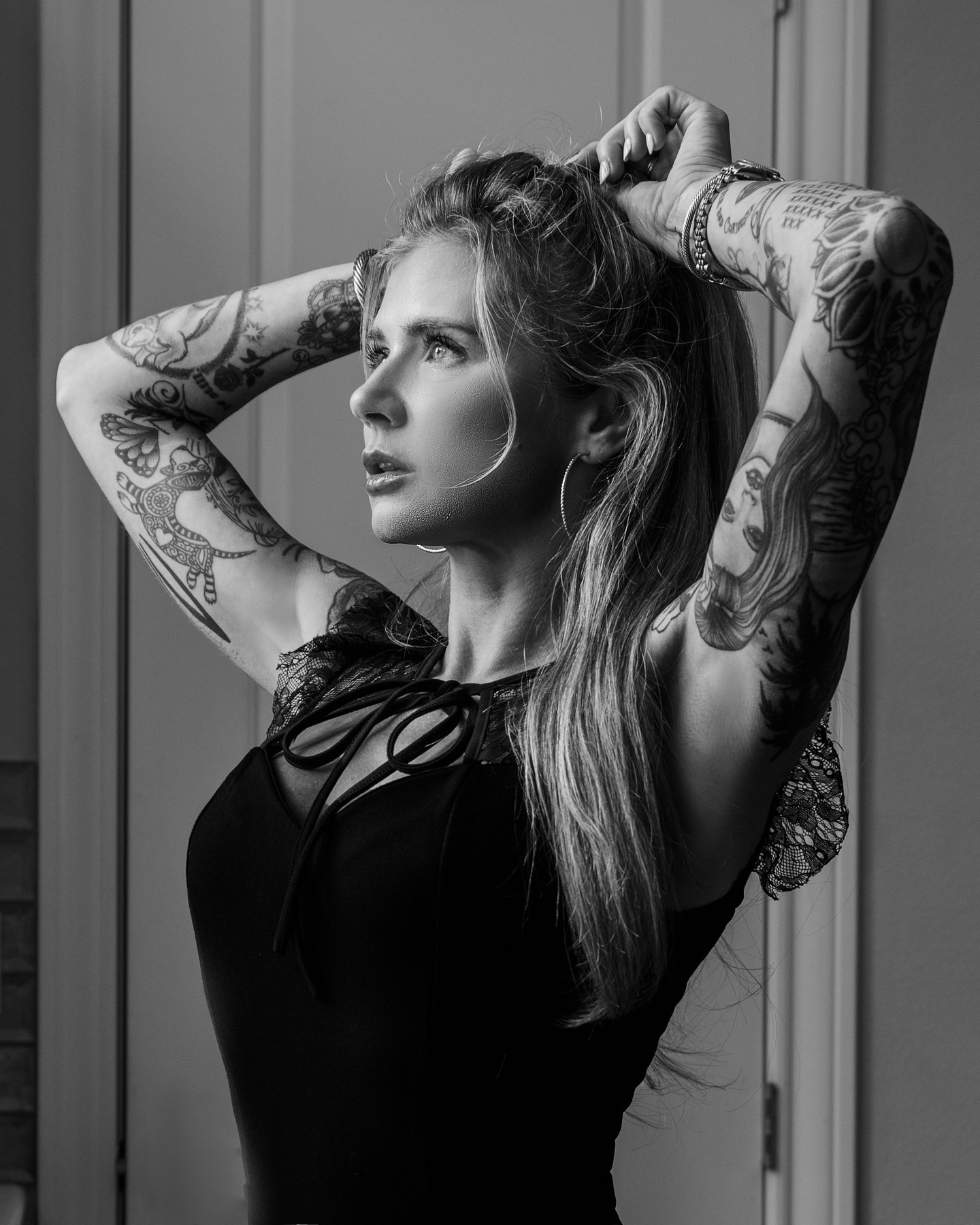 Black and white Austin boudoir portrait of a woman with tattooed arms raised in a black bodysuit by a window