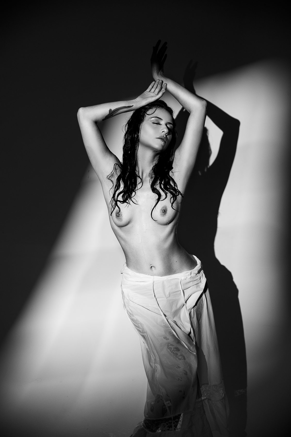 Black and white artistic nude portrait of a woman with her arms raised in window light and a sheer white skirt