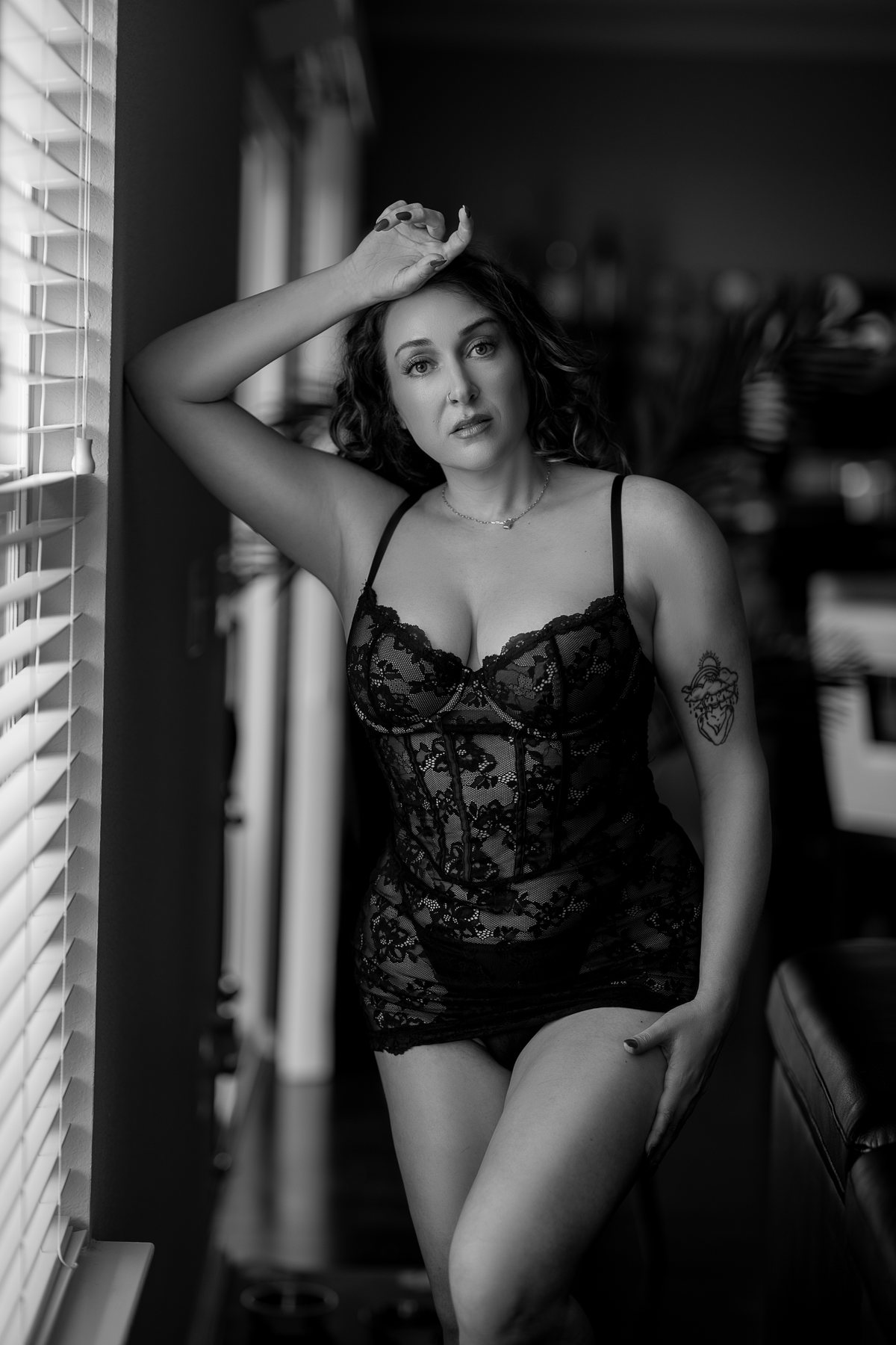 Black and white boudoir portrait of a woman in black lace lingerie beside window blinds in an Austin studio