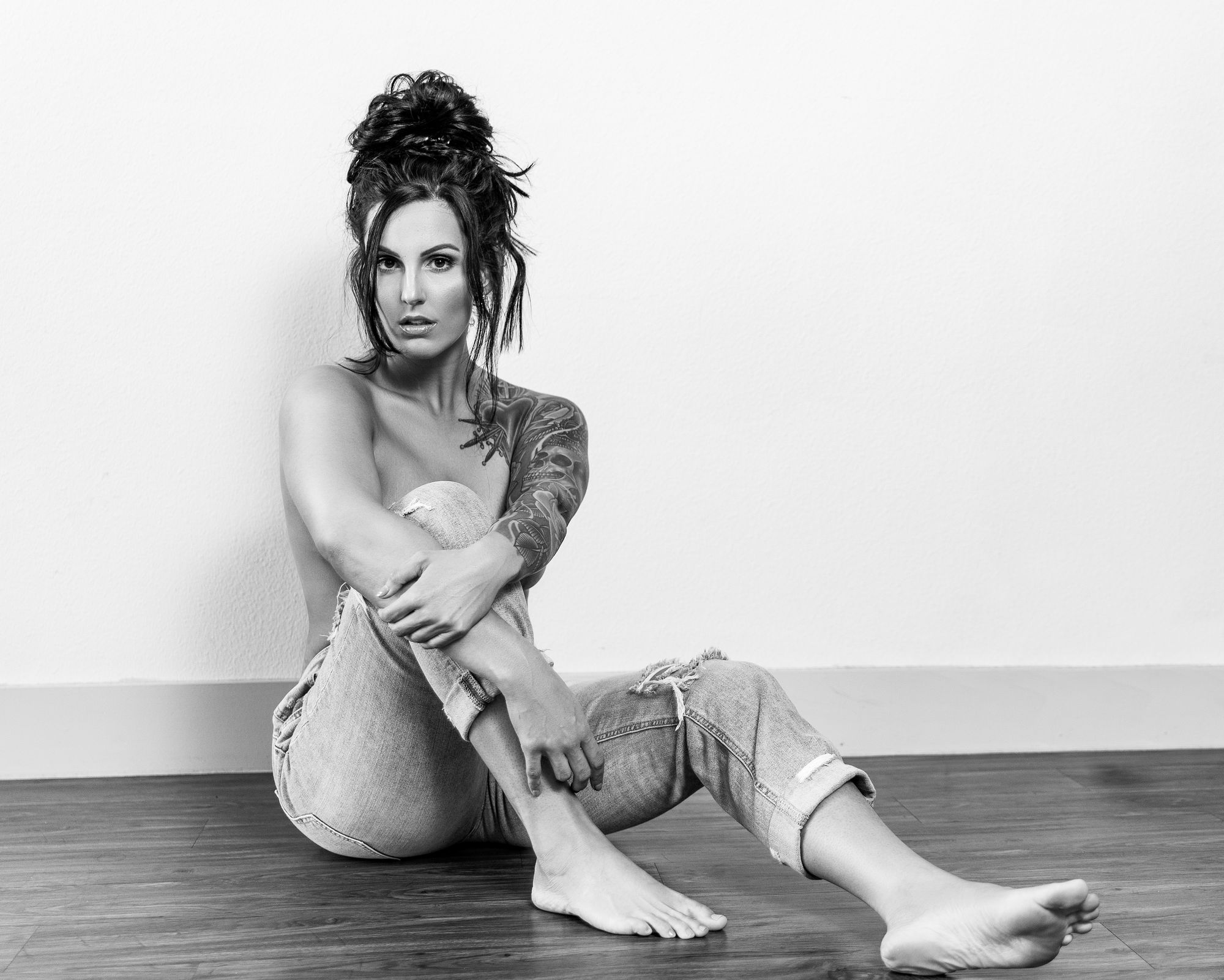 Black and white portrait of a woman seated on a wooden floor in jeans with her arms around her knees