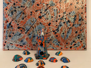Abstract orange-blue splatter painting behind a small painted pedestal and scattered colorful pebble-like pieces on a white surface