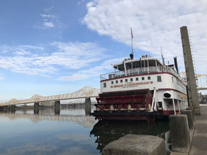 Paddle steamer "Belle of Louisville" docked on a calm river, with a bridge in the background and a clear blue sky. Tranquil scene.