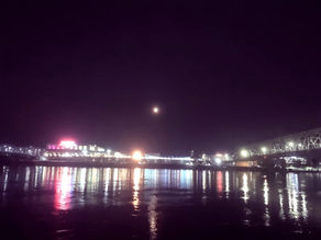 Night cityscape with bright neon lights reflecting on water, a glowing moon in the sky, and a bridge on the right. Calm and serene mood.