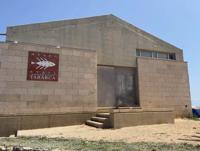 Beige building with "Museo Nueva Tabarca" sign featuring a fish logo. Steps lead to a metal door. Clear sky and sandy ground surround it.