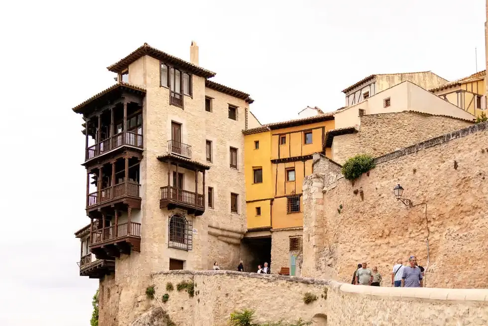 Stone buildings with wooden balconies on a cliffside, people walking below, cloudy sky, warm tones, historic atmosphere.