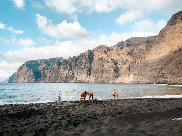 People enjoy a beach with black sand, a blue ocean, and towering cliffs in the background under a partly cloudy sky.