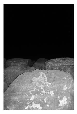 Black and white photograph of Aberavon.  Taken at night with coastal boulder sea defence boulders lit in the foreground and the blackness of the sea and beach at night in the background.