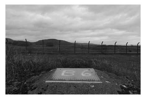 Black and white photograph in and around the former BP Baglan Bay.  A drain cover in the foreground, a fence in the middle and a hill and dark sky in the background.