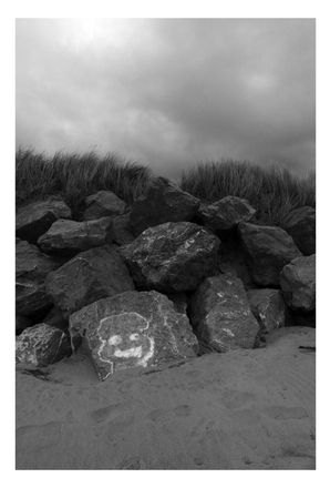 Black and white photograph at Crymlyn Burrows, South Wales.  Breakwater with a smiley face painted on one of the boulders.