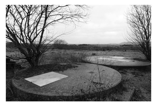 Black and white photograph in and around the former BP Baglan Bay.  A drain cover in the foreground covers and circular concrete shapes in the foreground.  A wintry landscape can be seen in the distance.