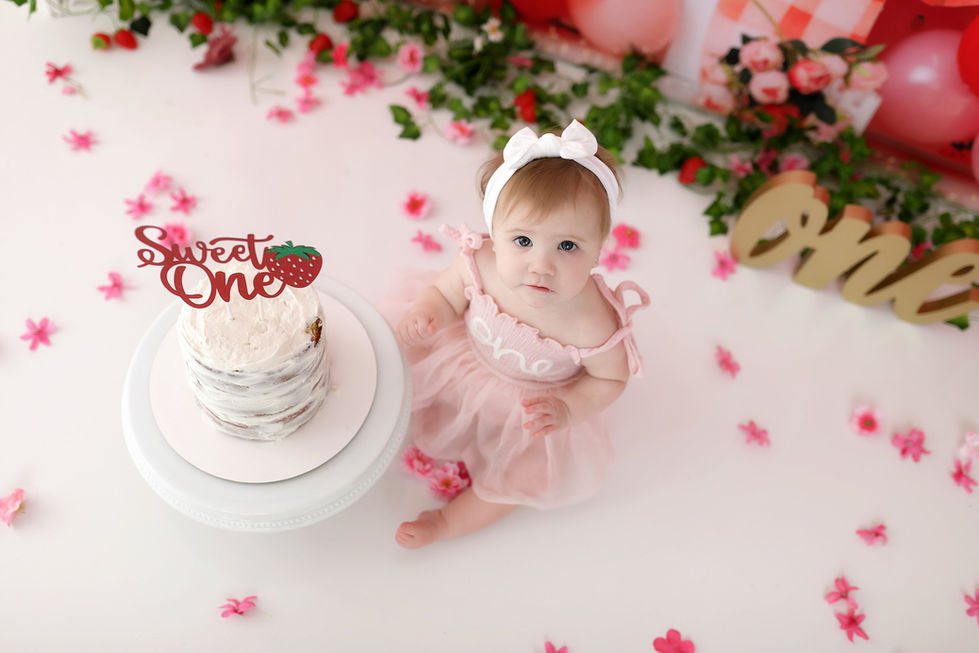 Strawberry-themed birthday backdrop with red and pink balloons, gingham bow, daisies, and strawberry props – ideal for cake smash and first birthday photography