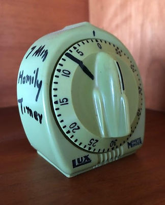 Whitish kitchen-style timer with markings for each minute. A large hand-marked line is at minute 7. The side of the timer has "7 Min Homily Timer" written on it.