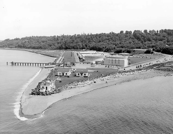 A triangular spit of land extending into a body of water, small lighthouse at its tip, other buildings behind, and a pier on the left. Behind the developed area is a large expanse of trees.