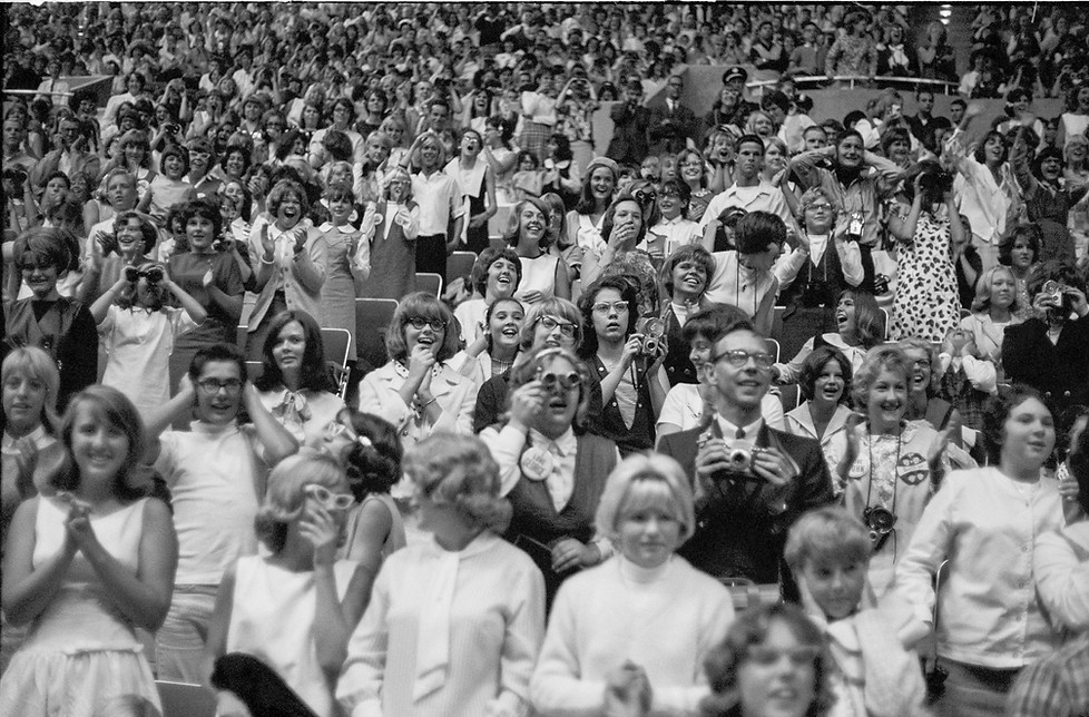 Black-and-white photo of a large crowd of people in auditorium seating; most standing up, some holding their hands over their ears, and some using binoculars.
