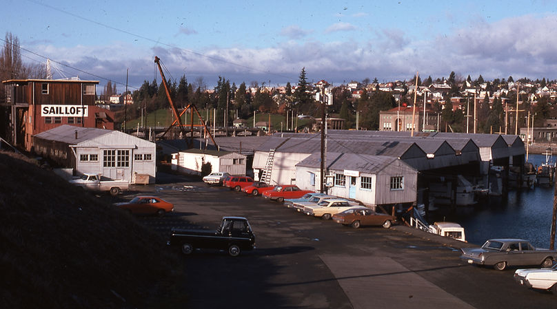 A series of about ten short buildings along a dock, water to the right, and cars parked to the left. Trees and more buildings are in the background. There is a light blue sky and a layer of low, puffy, light gray clouds.