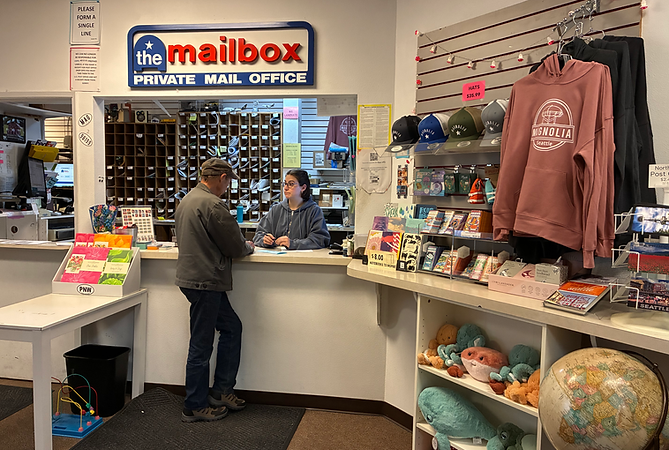 A mail service desk has an employee with shoulder-length dark hair and glasses behind it. A customer with a ballcap stands on the other side. The desk is surrounded by a colorful array of apparel, toys, cards, and other gifts.