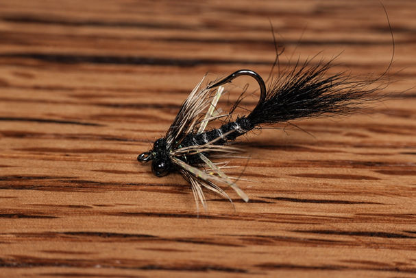 Black fly with dark bead-chain eyes, hackle, pale rubber legs, and an upright hook point resting on a warm wooden surface.