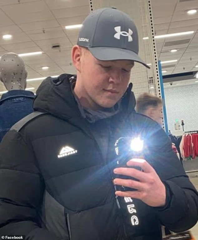 Man in gray Under Armour cap and black jacket takes a mirror selfie in a store, camera flash on, with clothing racks behind.