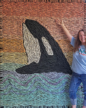 Rebecca Hoooper standing next to a 7 foot tall weaving of a killer whale breaching in the ocean with a sunset background