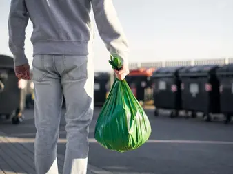 Imagem de uma pessoa segurando um saco de lixo em frente a contentores de resíduos