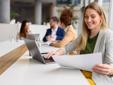 Business woman with blonde hair in foreground, working on computer and looking at paper. Three colleagues in the background strategising.