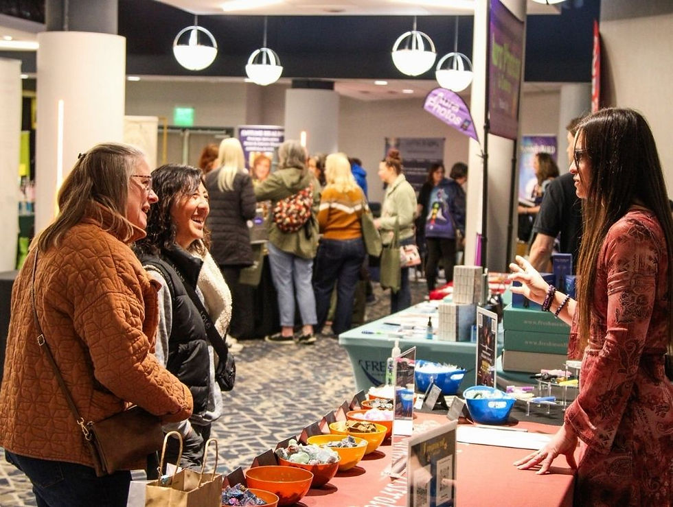 Women interacting at a vendor booth with colorful bowls in a busy indoor market. Bright overhead lights; lively, conversational mood.