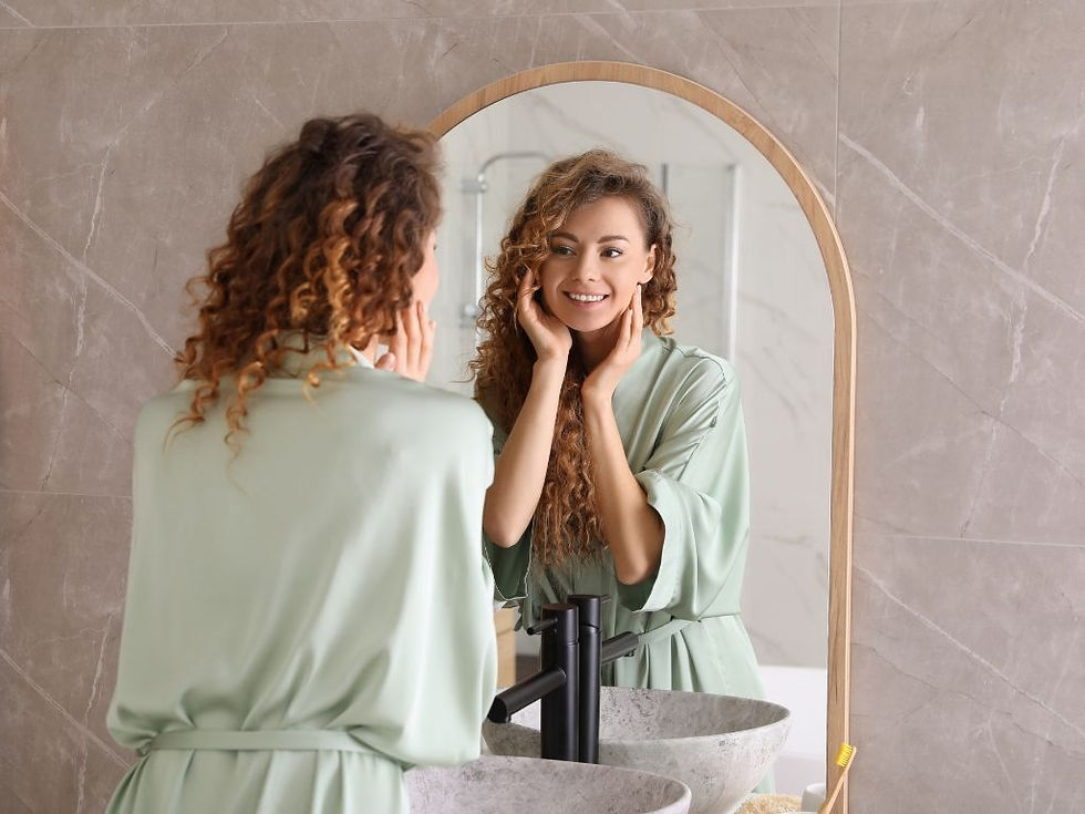 Woman in a mint robe smiles at her reflection, touching her cheeks in a modern bathroom with a round mirror.