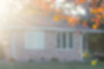 Sunlit brick house with white windows, autumn leaves in foreground, and a yellow toy truck on the porch.