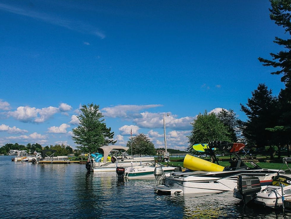 Boats docked on a calm lake under a bright blue sky with fluffy clouds. Green trees line the shore, creating a peaceful atmosphere.