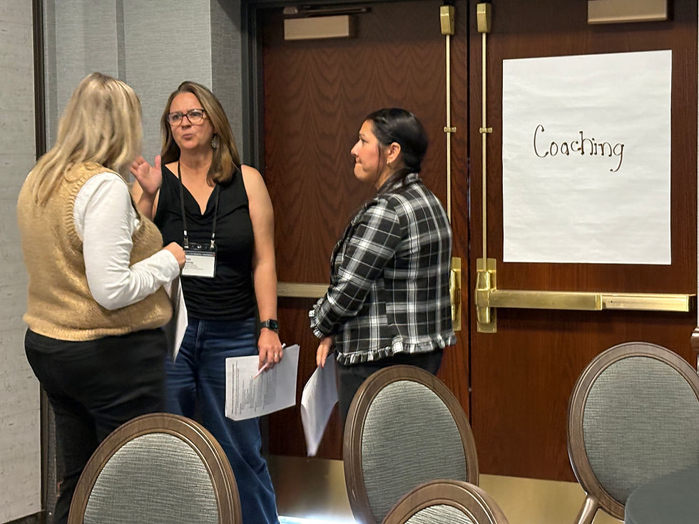 Three women talking by door with paper noting "Coming" and Real Leaders Real Impact