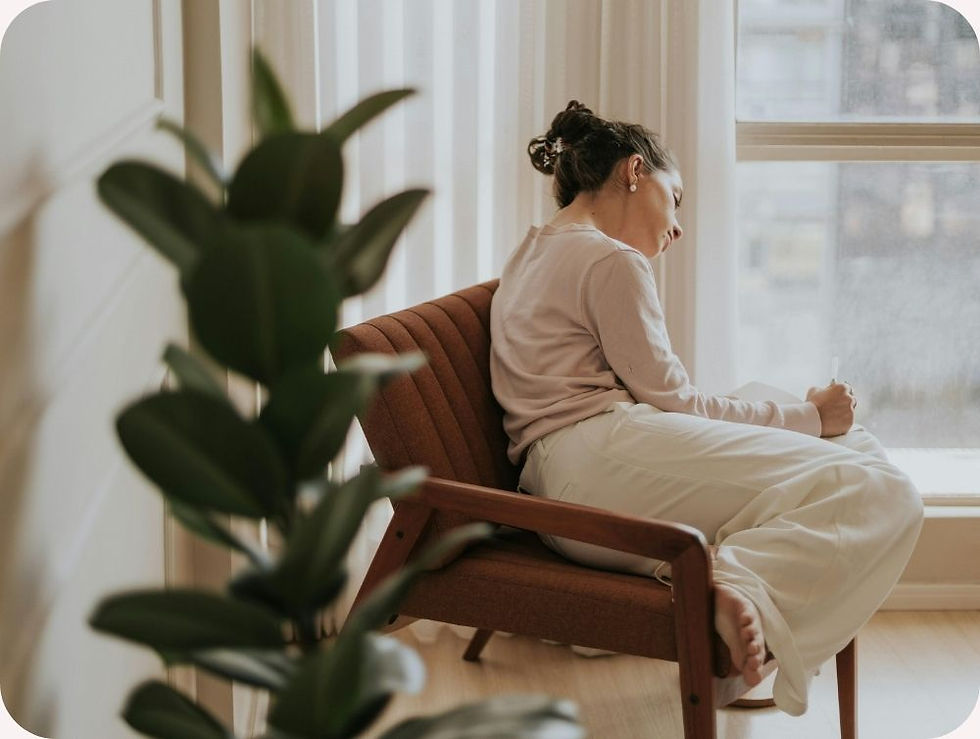 Woman in a beige sweater and white pants writes in a notebook while reclining on a brown chair near a window. Green plant in the foreground.